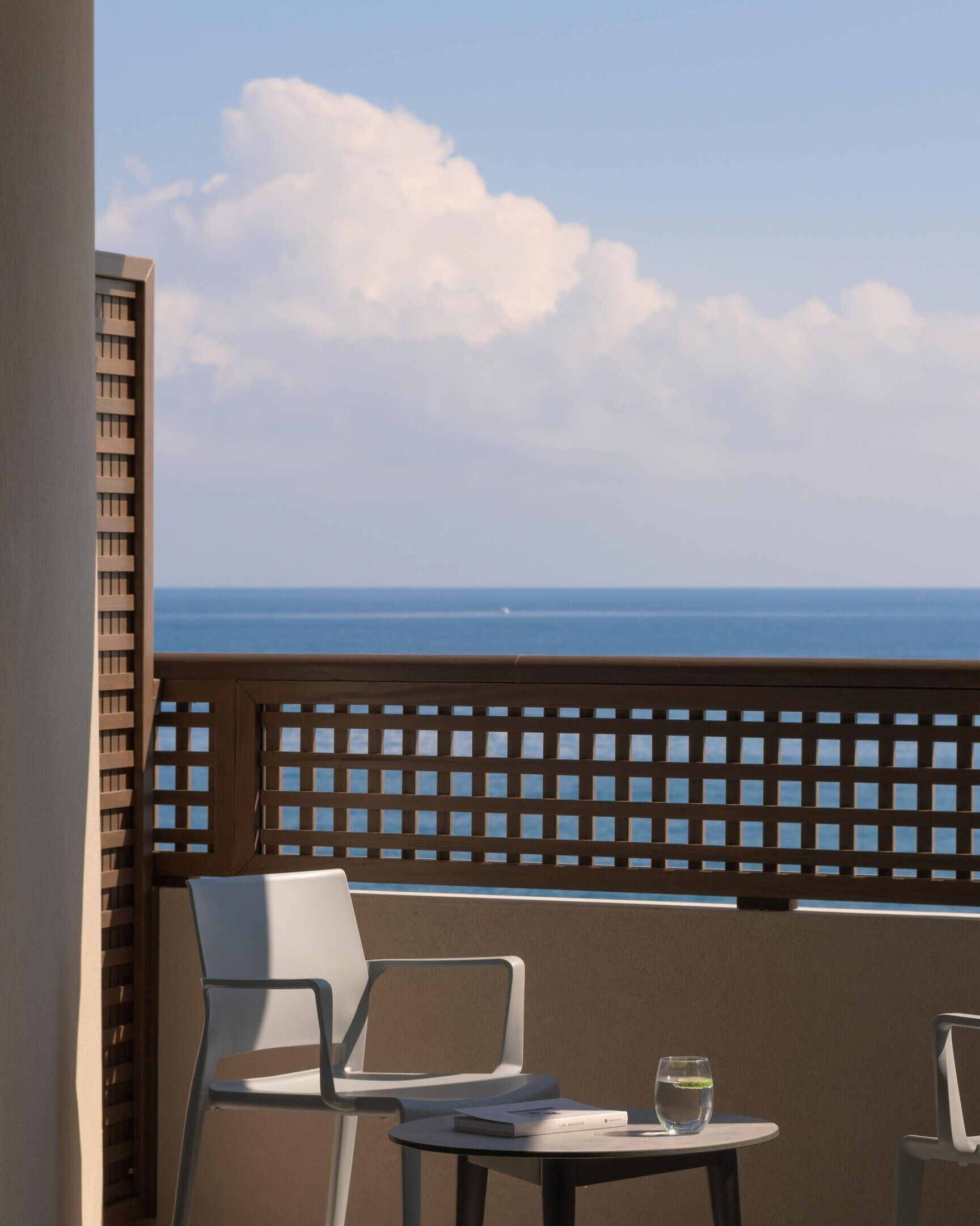 A balcony with a view of the ocean, a white chair, a small round table, and a glass of water.