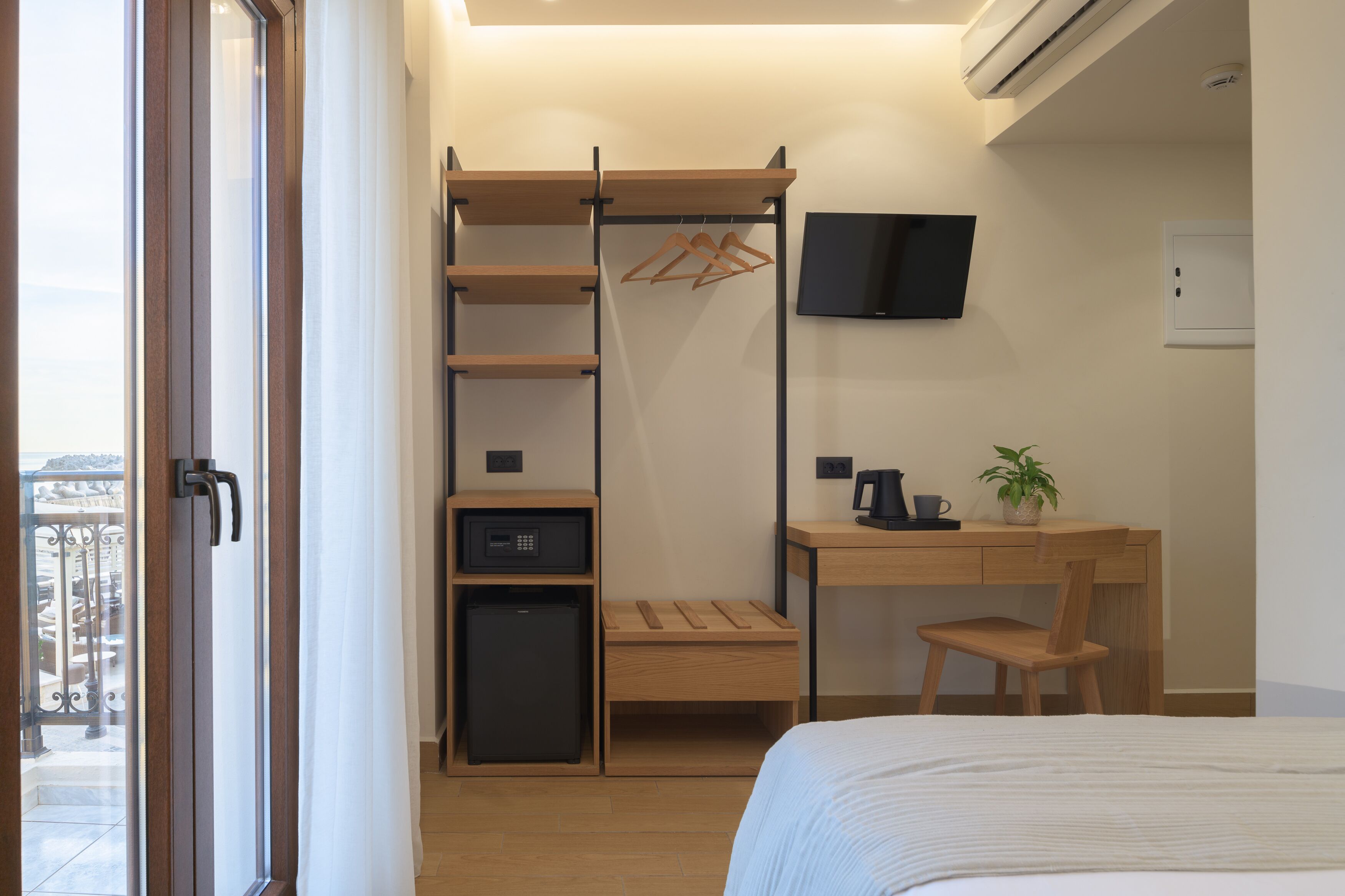 Habitación de hotel compacta con cama, escritorio con silla, refrigerador pequeño, televisor montado en la pared y armario de madera con perchero.