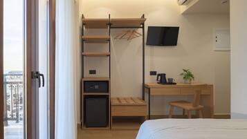 Habitación de hotel compacta con cama, escritorio con silla, refrigerador pequeño, televisor montado en la pared y armario de madera con perchero.