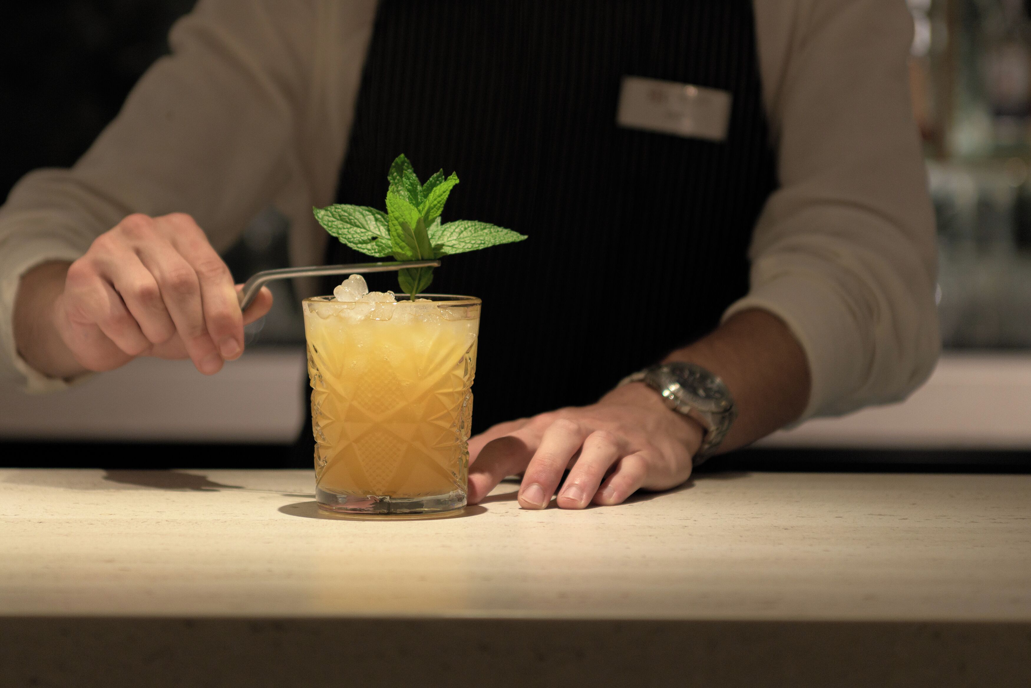 A person in a white shirt and black apron preparing a drink with a mint garnish.