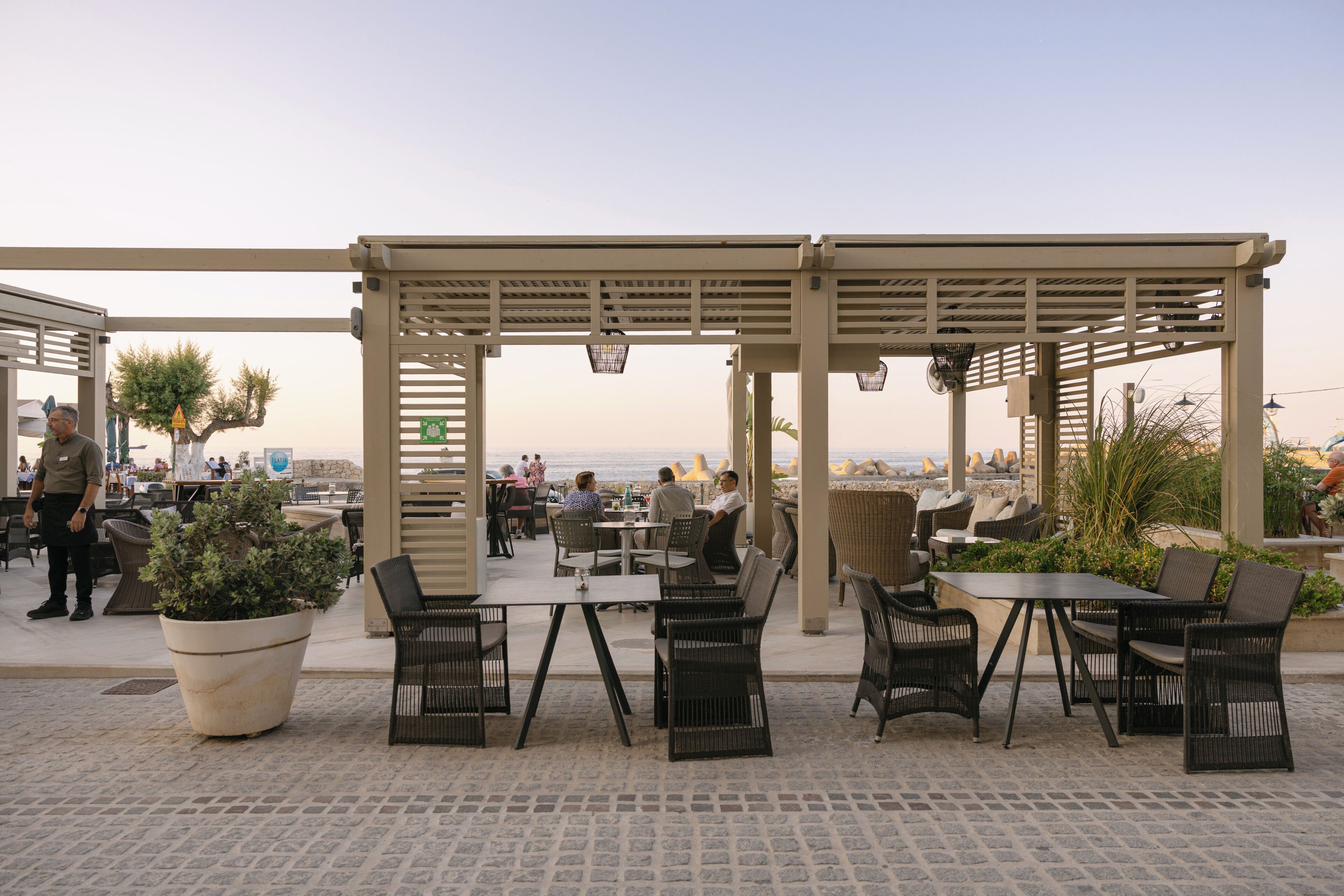 An outdoor cafe with tables and chairs, a large potted plant, and a view of the sea.