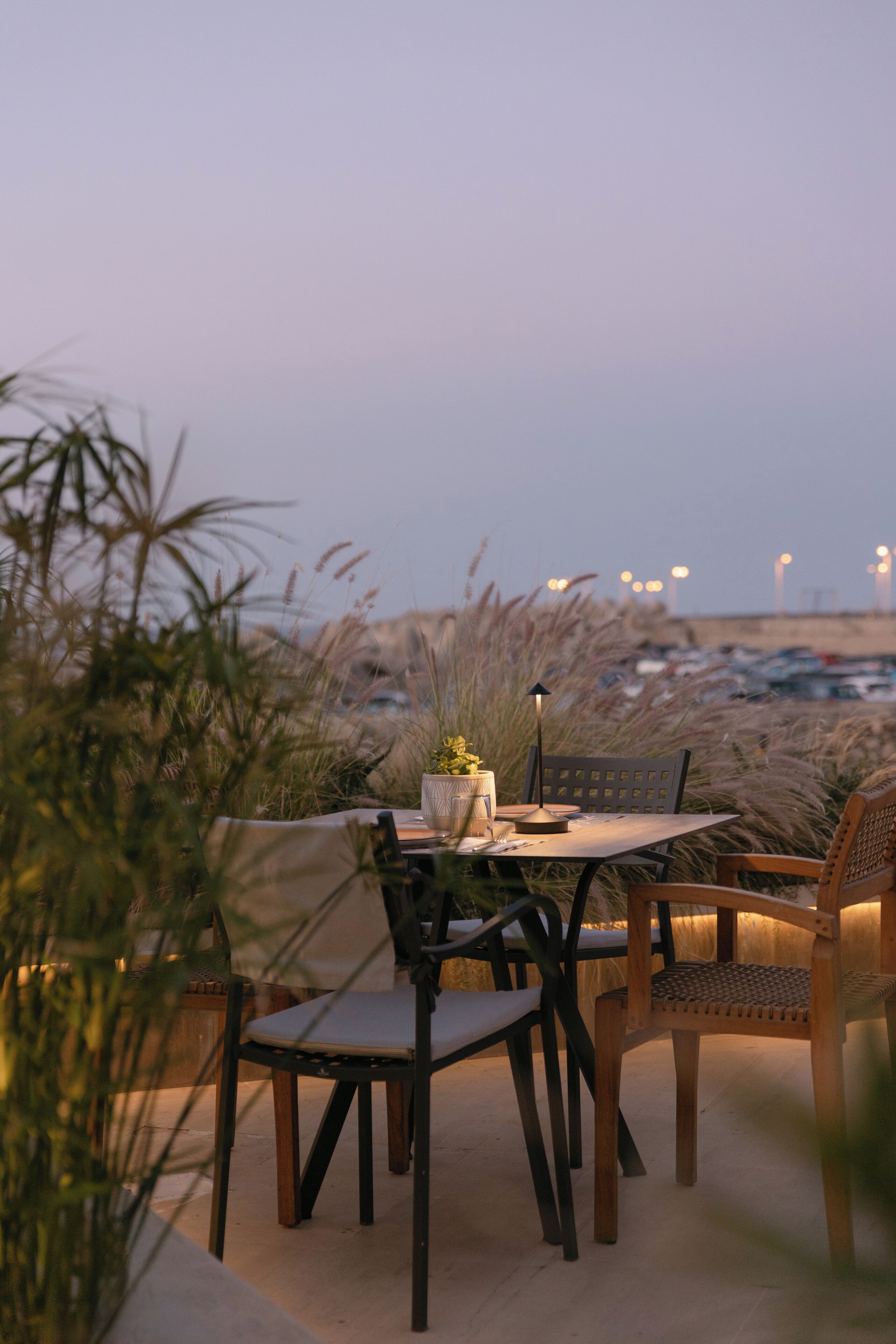 An outdoor dining area with a table and chairs, surrounded by tall grasses and a view of a parking lot in the background.