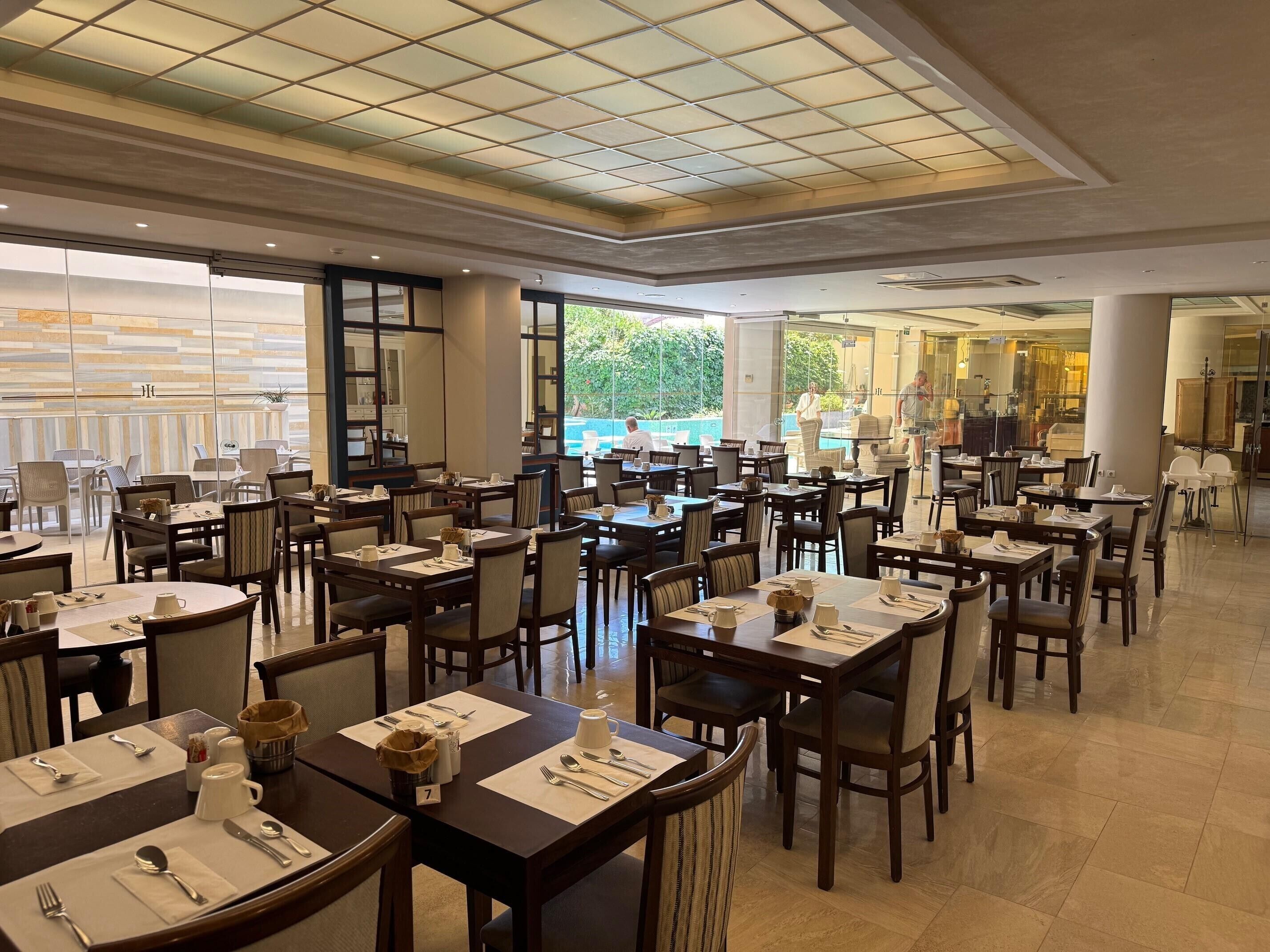 A spacious dining area with multiple tables and chairs, large windows offering a view of the outdoors, and a modern ceiling design.