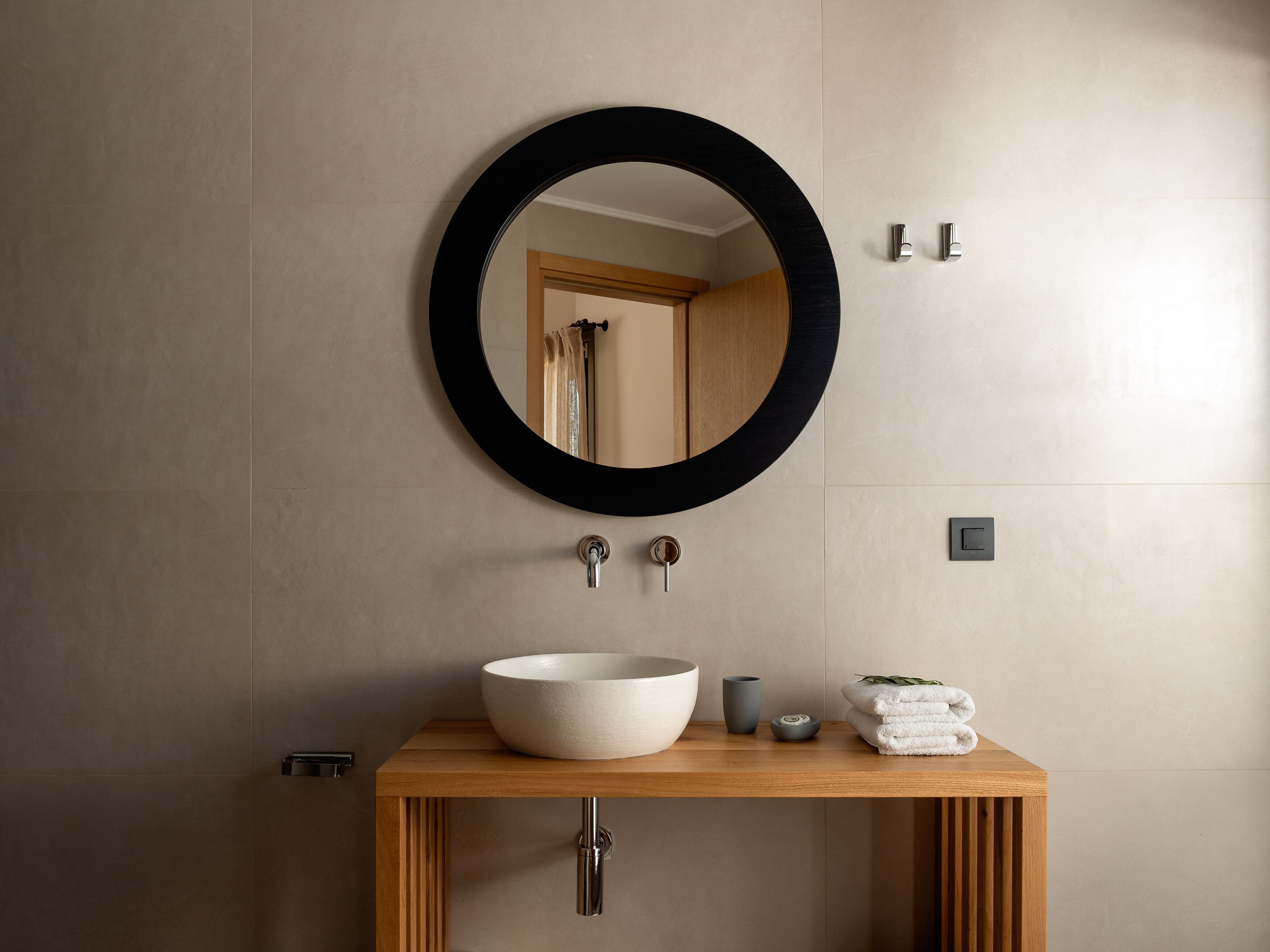 Un baño con un espejo redondo, un tocador de madera, un lavabo blanco, un jarrón pequeño y una pila de toallas.