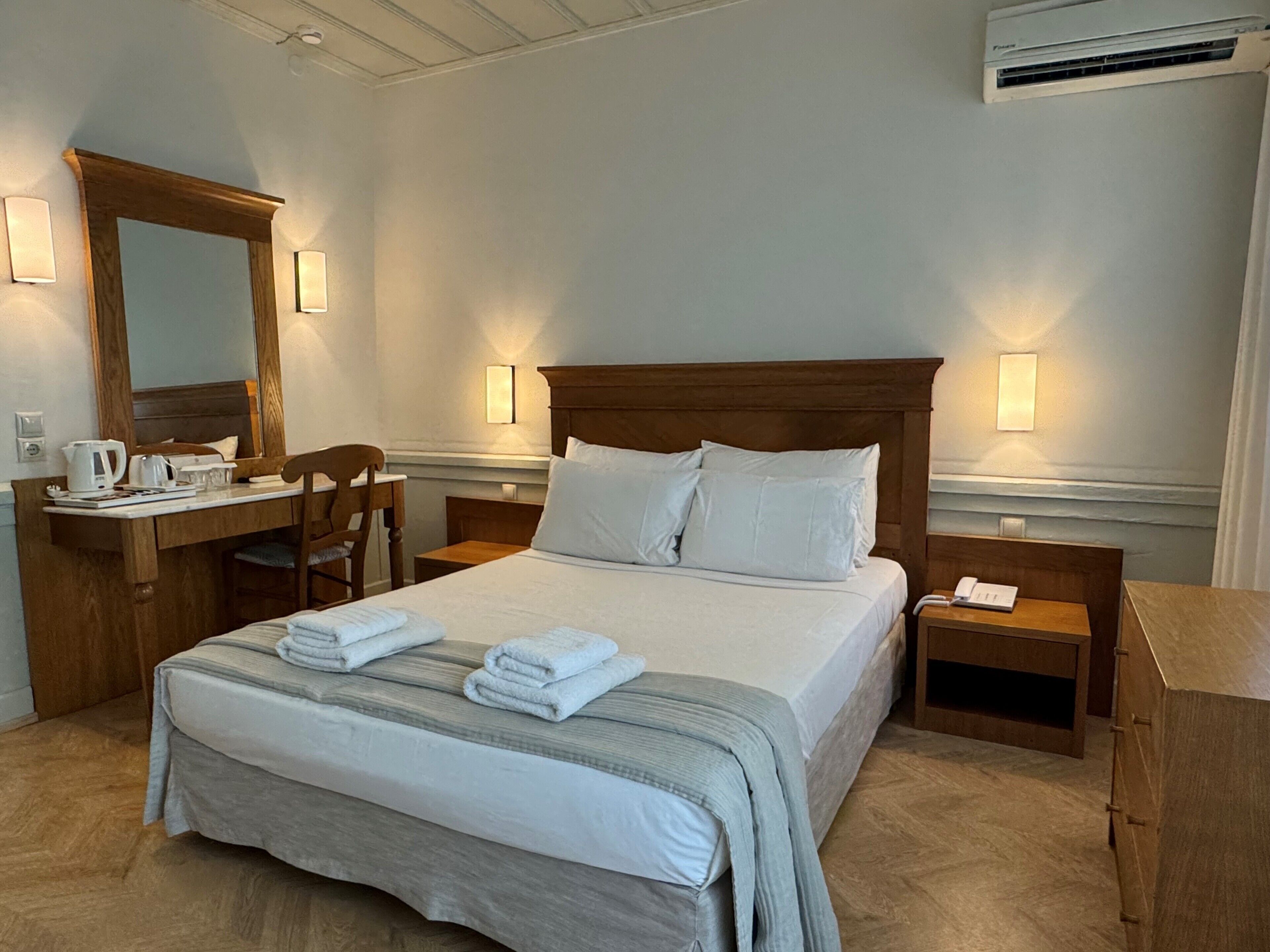 A hotel room with a large bed, a wooden desk with a chair, a mirror, and bedside tables with lamps.
