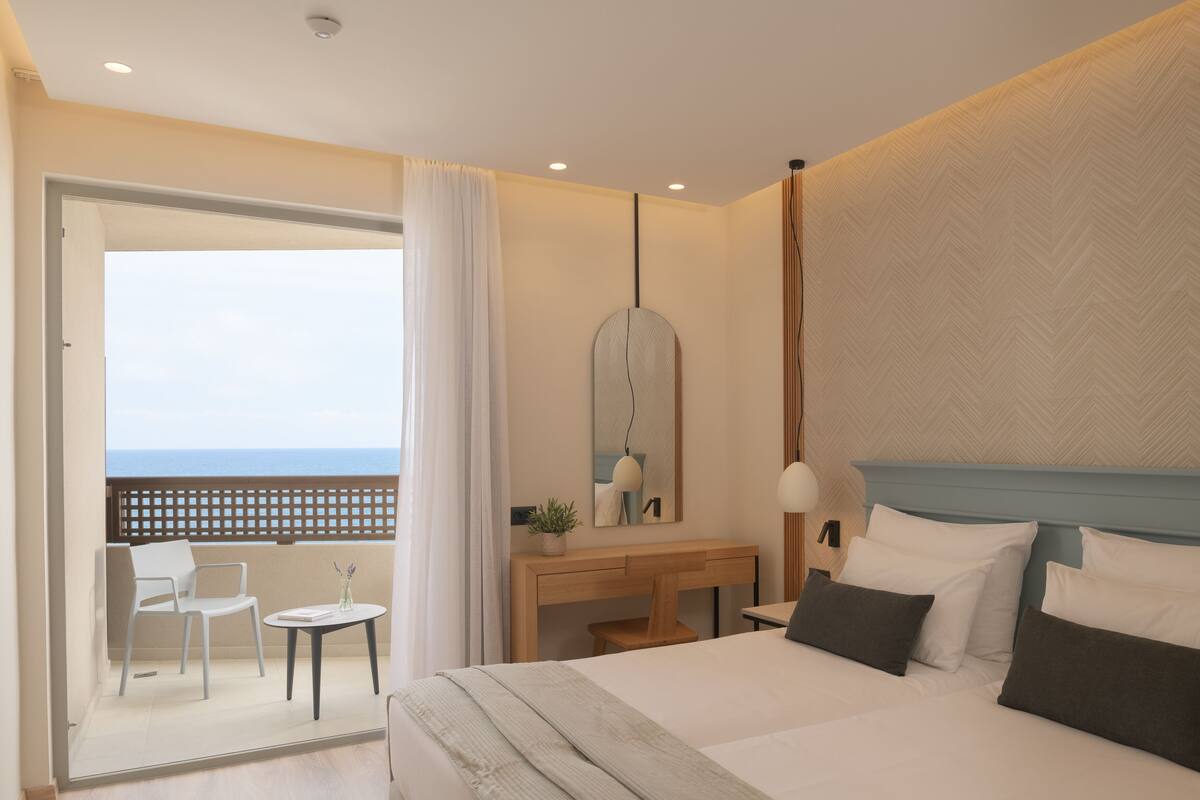 Quarto de hotel com uma cama, uma escrivaninha, uma cadeira, uma mesinha e vista para o mar através de uma porta de varanda.