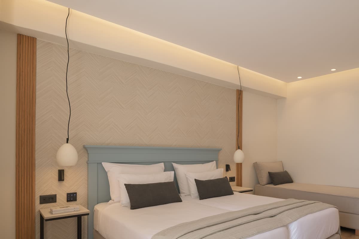 Quarto de hotel moderno com uma cama grande, uma mesa de cabeceira, um sofá e uma luminária fixada na parede.