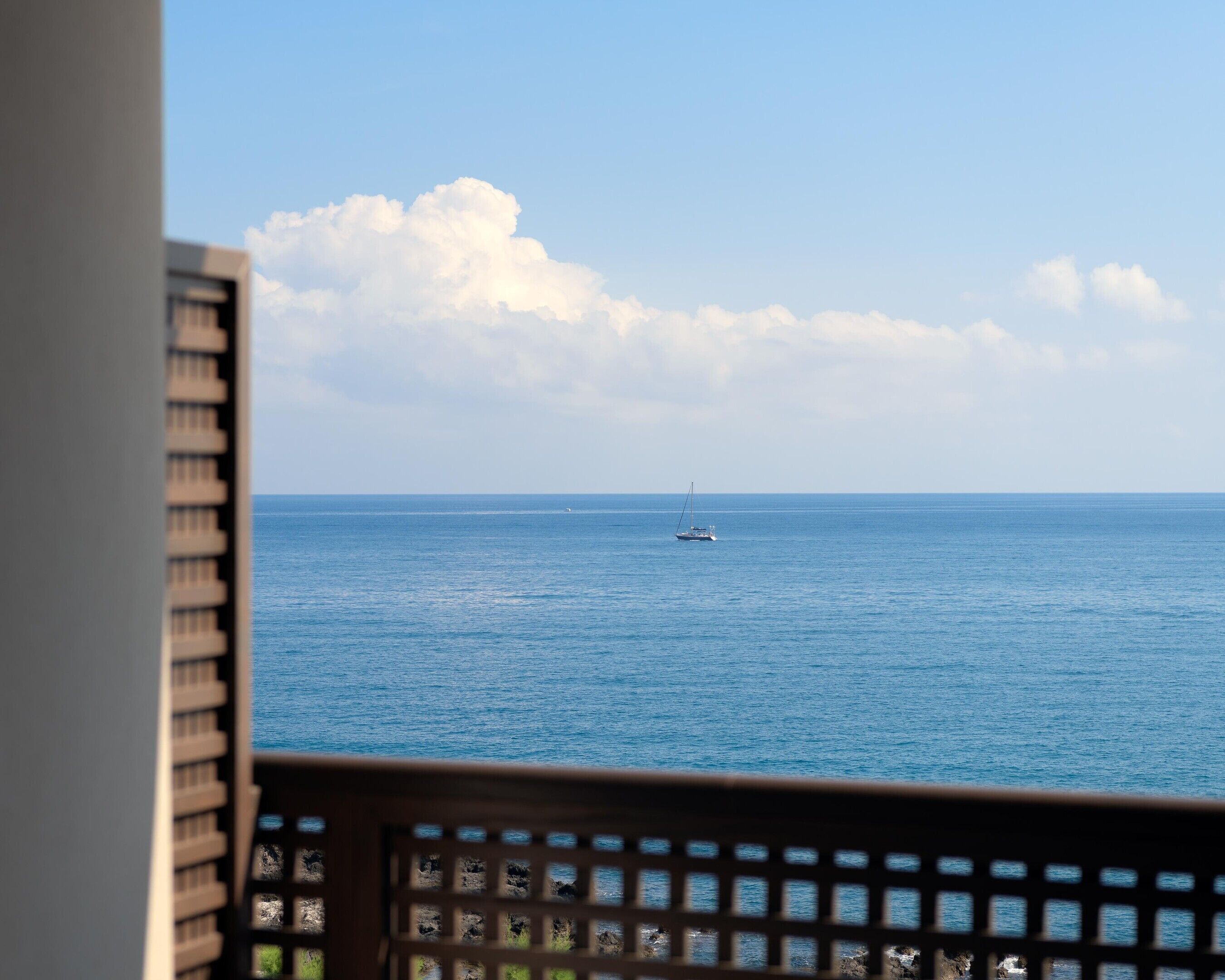 Vista del océano con un velero a lo lejos, visible a través de una ventana con celosía de madera.