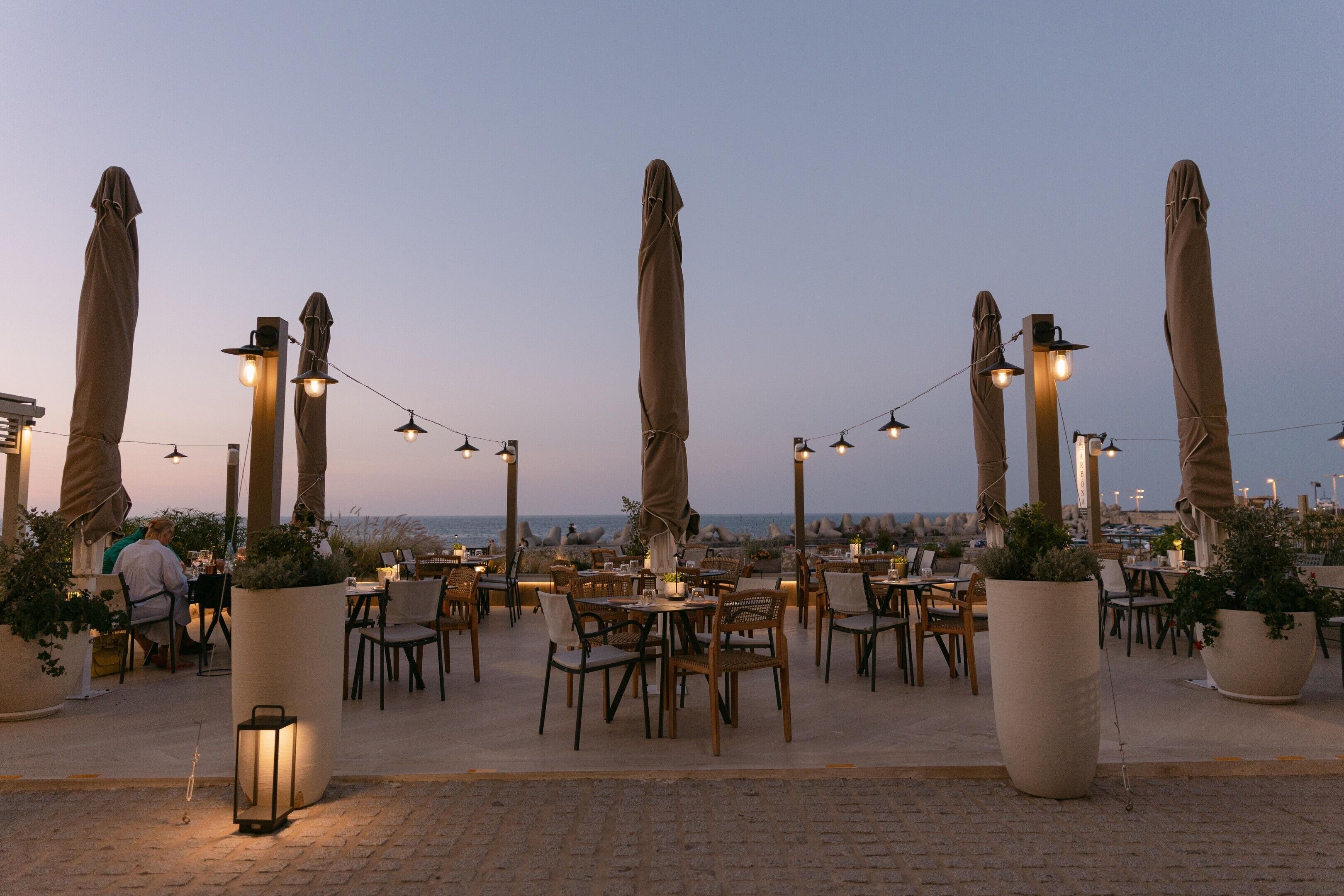 Un espacio de comedor al aire libre con mesas, sillas, sombrillas y luces colgantes.