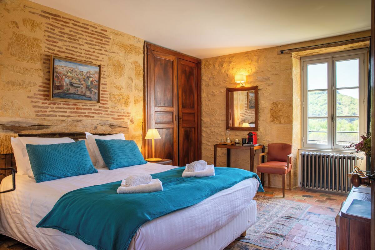 A bedroom with a large bed, a wooden door, a small table with a lamp, a chair, and a window with a view of greenery.