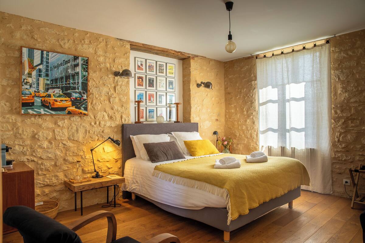 A cozy bedroom with a stone wall, a bed with a yellow blanket, a nightstand with a lamp, and a framed cityscape picture above the bed.