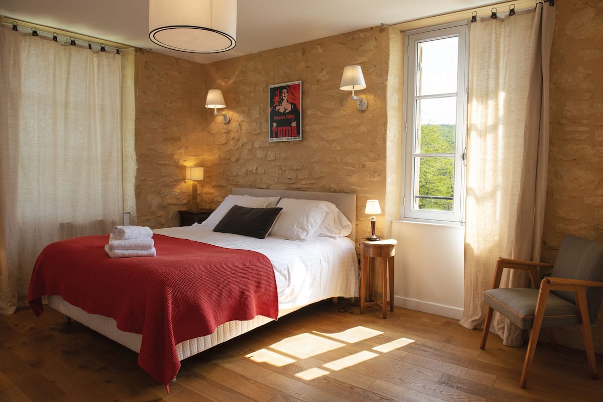 A bedroom with a bed, a nightstand, a chair, a window, and a stone wall.