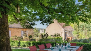 Un área de comedor al aire libre con una mesa preparada para cuatro personas, rodeada de follaje y un árbol.