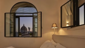 Une chambre d’hôtel avec un lit, une fenêtre donnant sur un bâtiment historique, une lampe de chevet et un miroir au mur.
