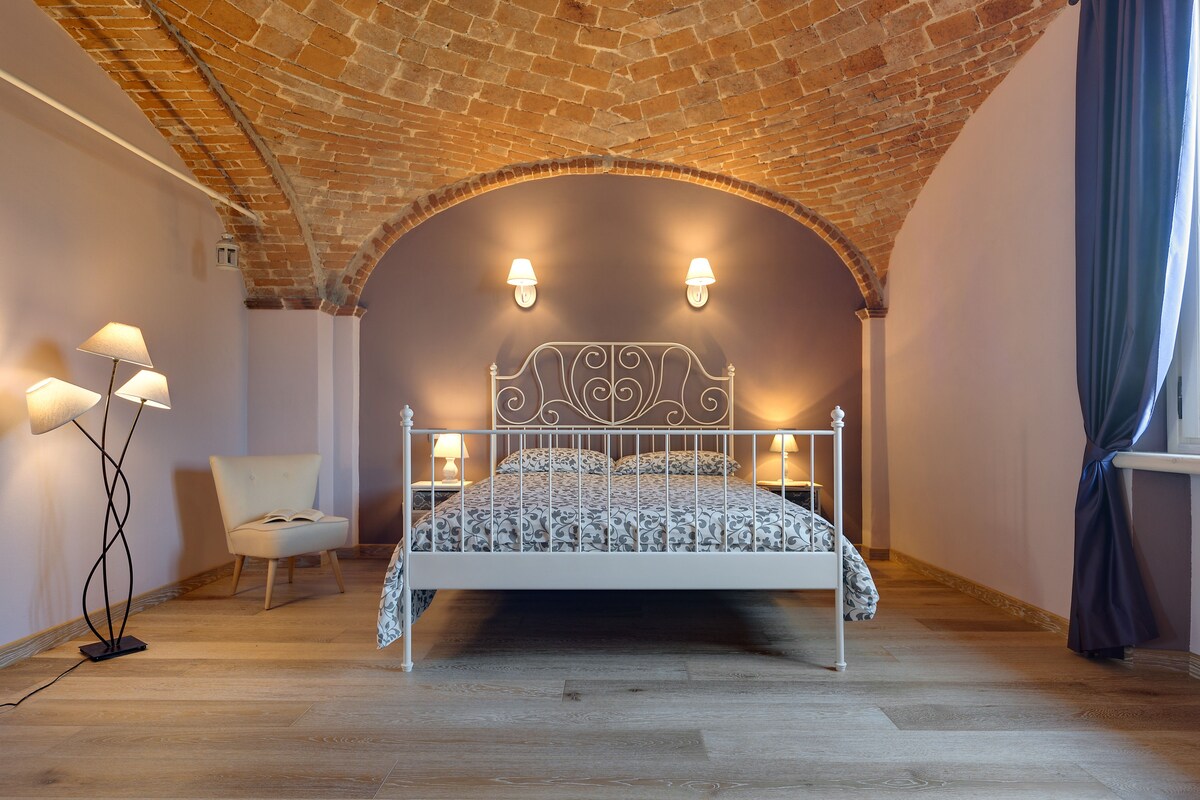 Camera da letto con soffitto in mattoni, struttura del letto in metallo bianco, biancheria a motivi decorativi e due lampade a parete.