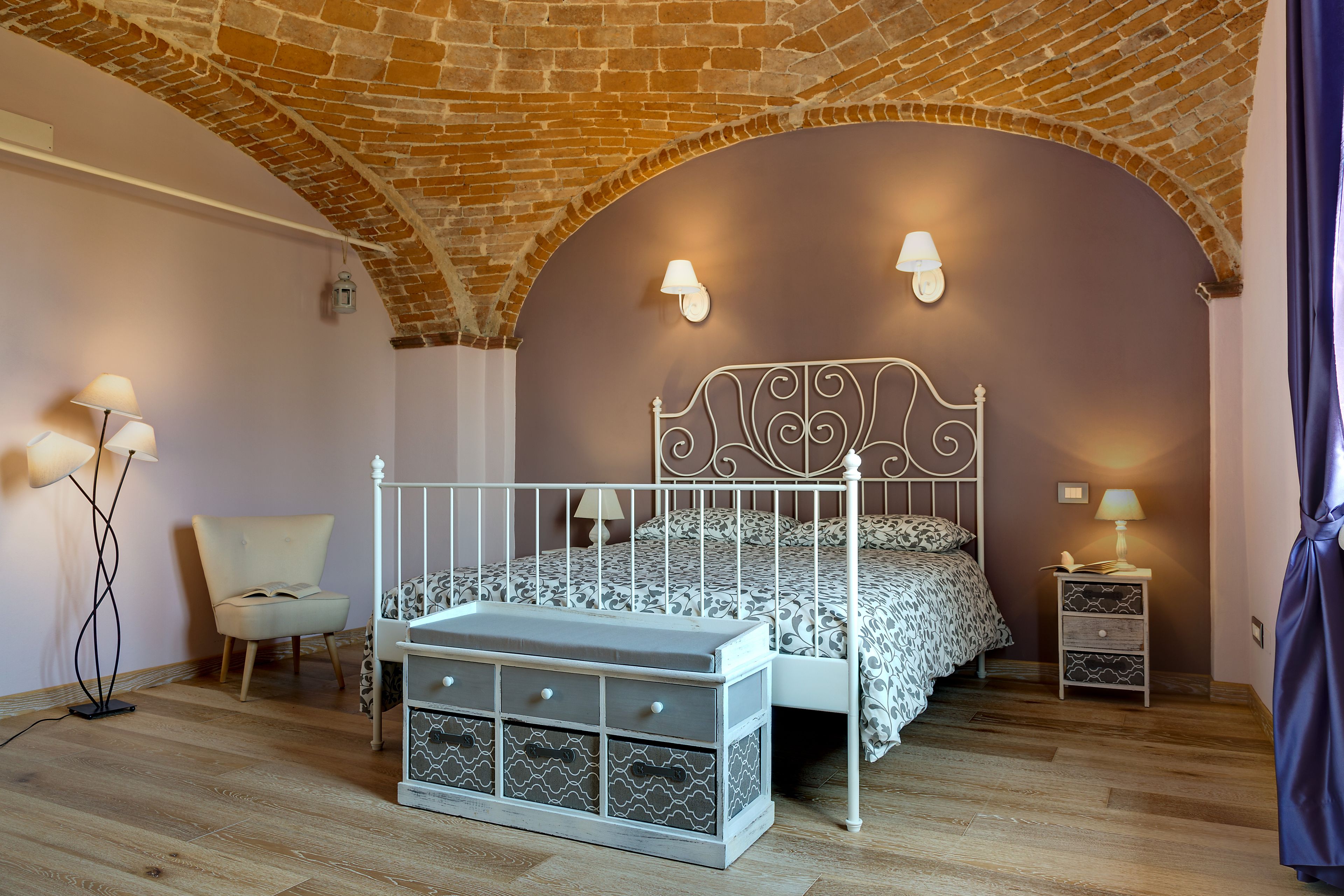 Une chambre avec un plafond en briques, un cadre de lit en métal blanc, une literie à motifs et une table de chevet avec des tiroirs.
