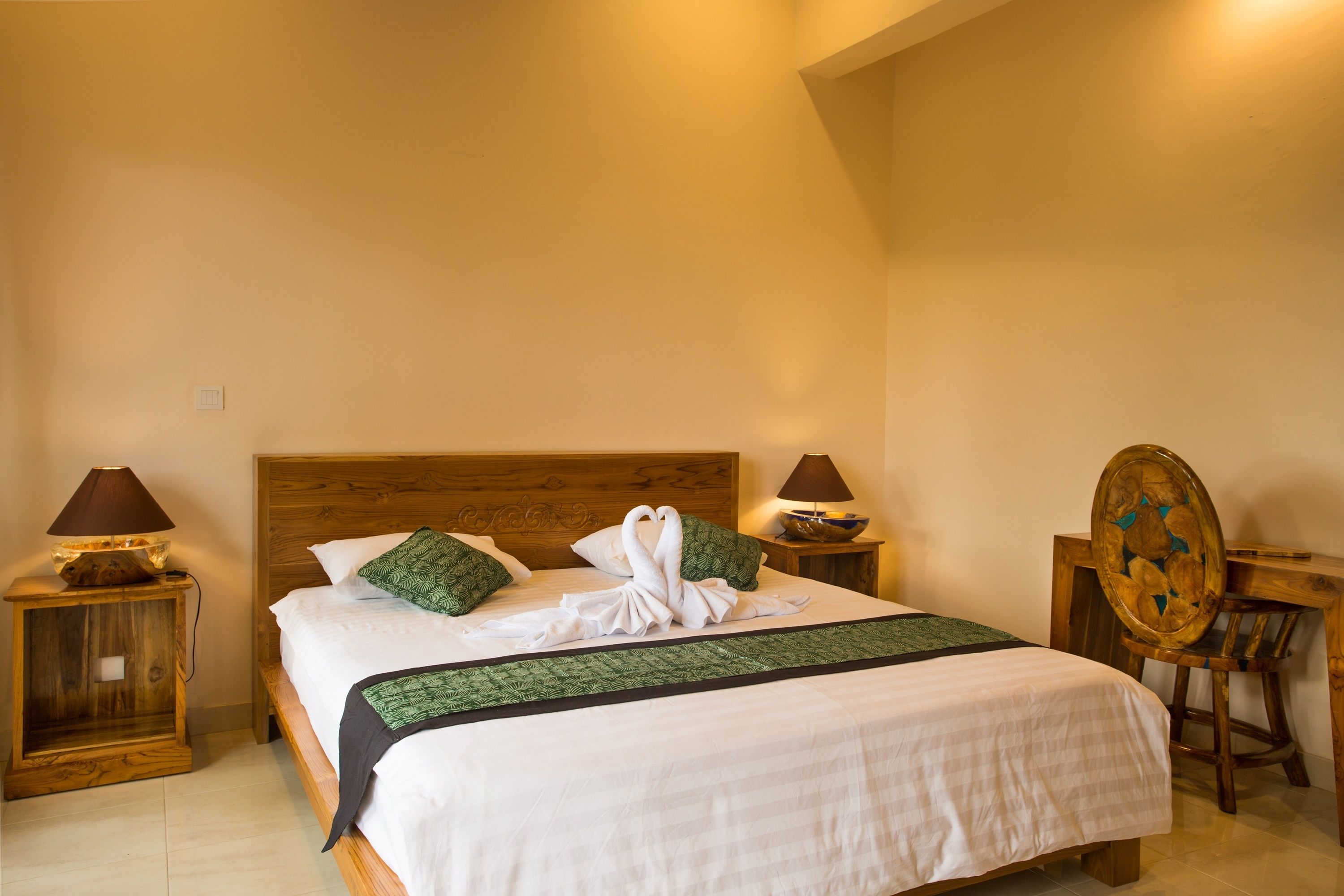 Una cama bien hecha con cabecera de madera, dos mesitas de noche con lámparas, una silla de madera y una mesita pequeña con un cuenco.