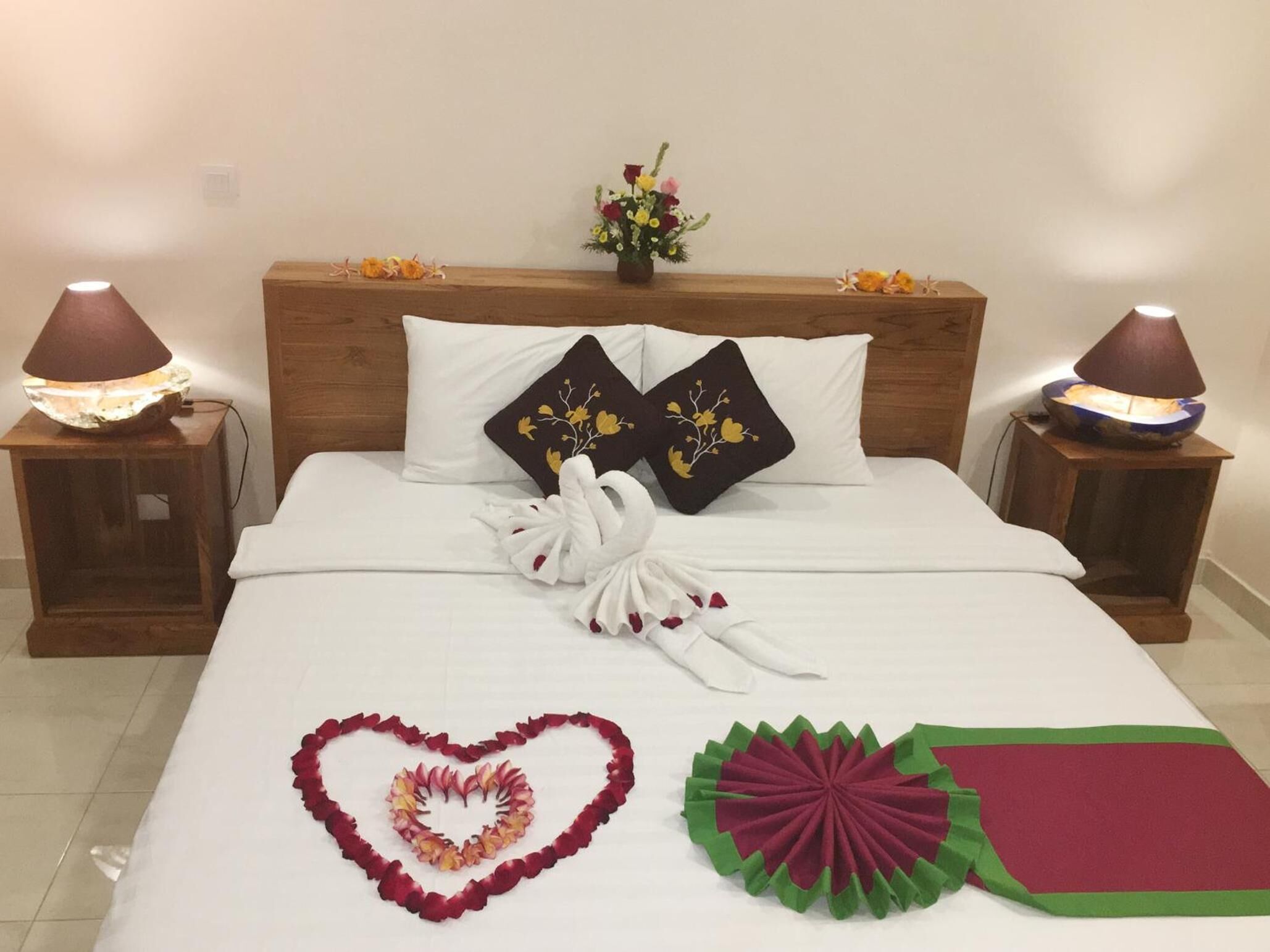 A neatly made bed with a heart-shaped flower arrangement, two decorative pillows, and a small vase of flowers on the headboard.