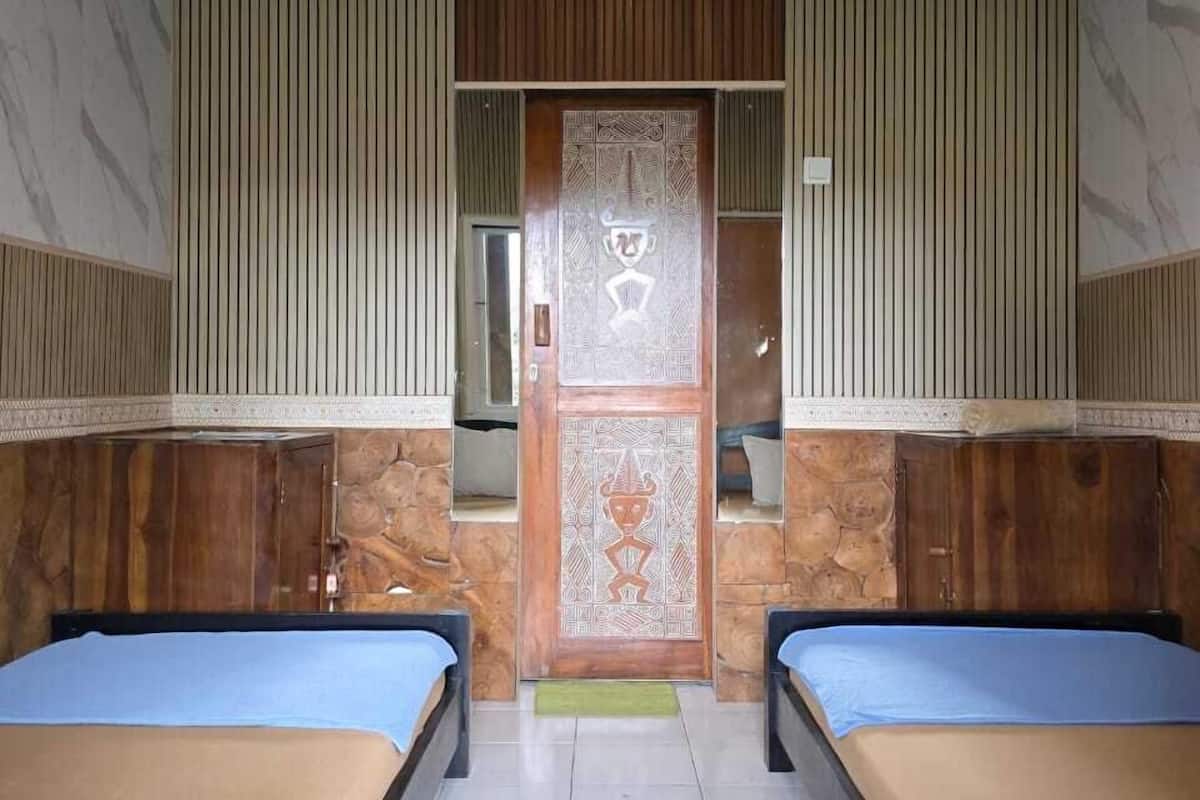 Quarto com duas camas, porta de madeira com detalhes entalhados, ventilador de teto e parede de pedra.