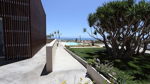 Un edificio moderno con un sendero de hormigón que lleva a una zona de piscina, rodeada de vegetación y vistas al océano.