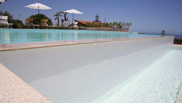 Piscina con sillones de descanso y sombrillas, rodeada de jardines con árboles y vistas al océano.