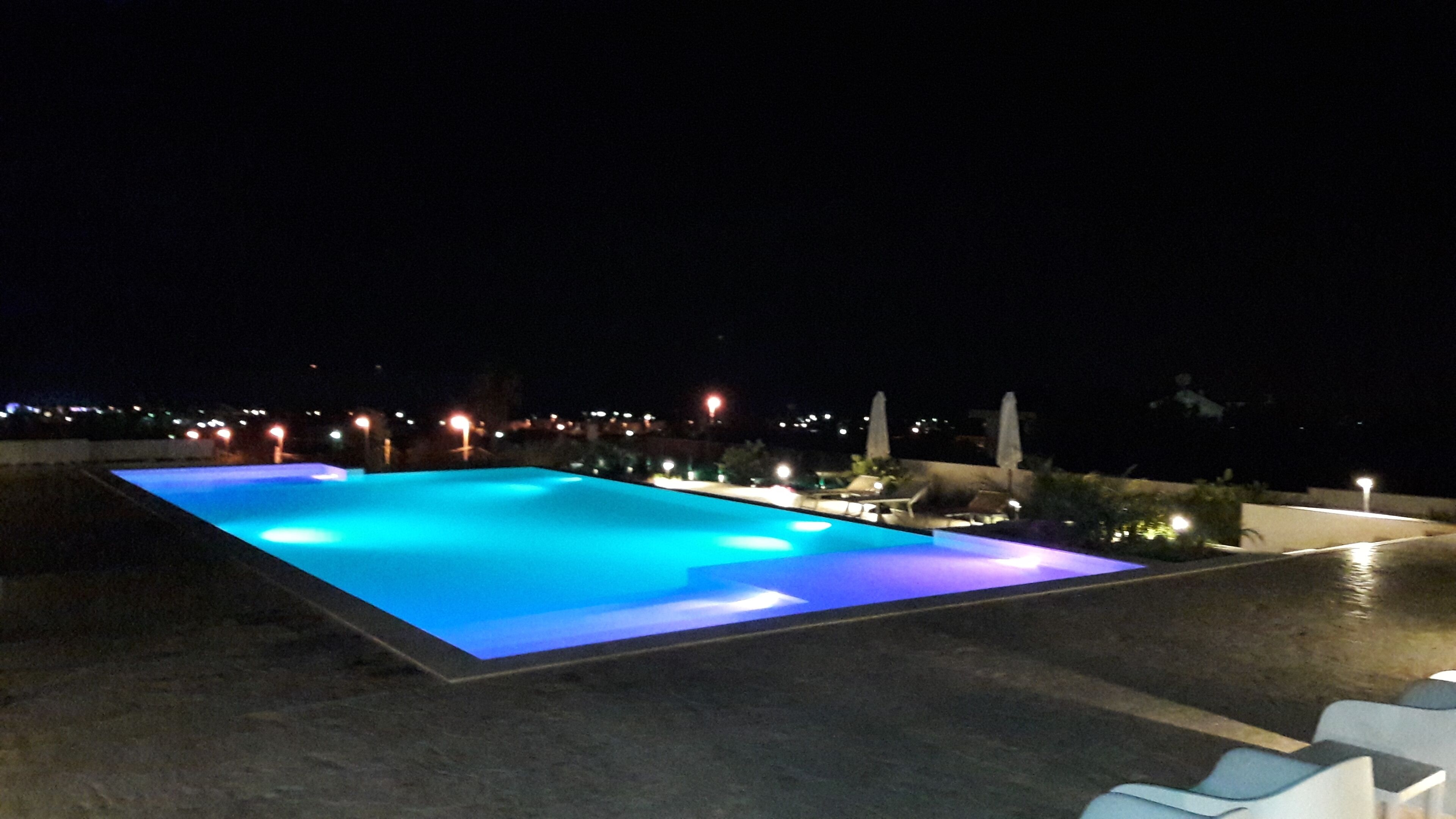 Piscina retangular com bordas iluminadas, rodeada por espreguiçadeiras e um muro baixo.
