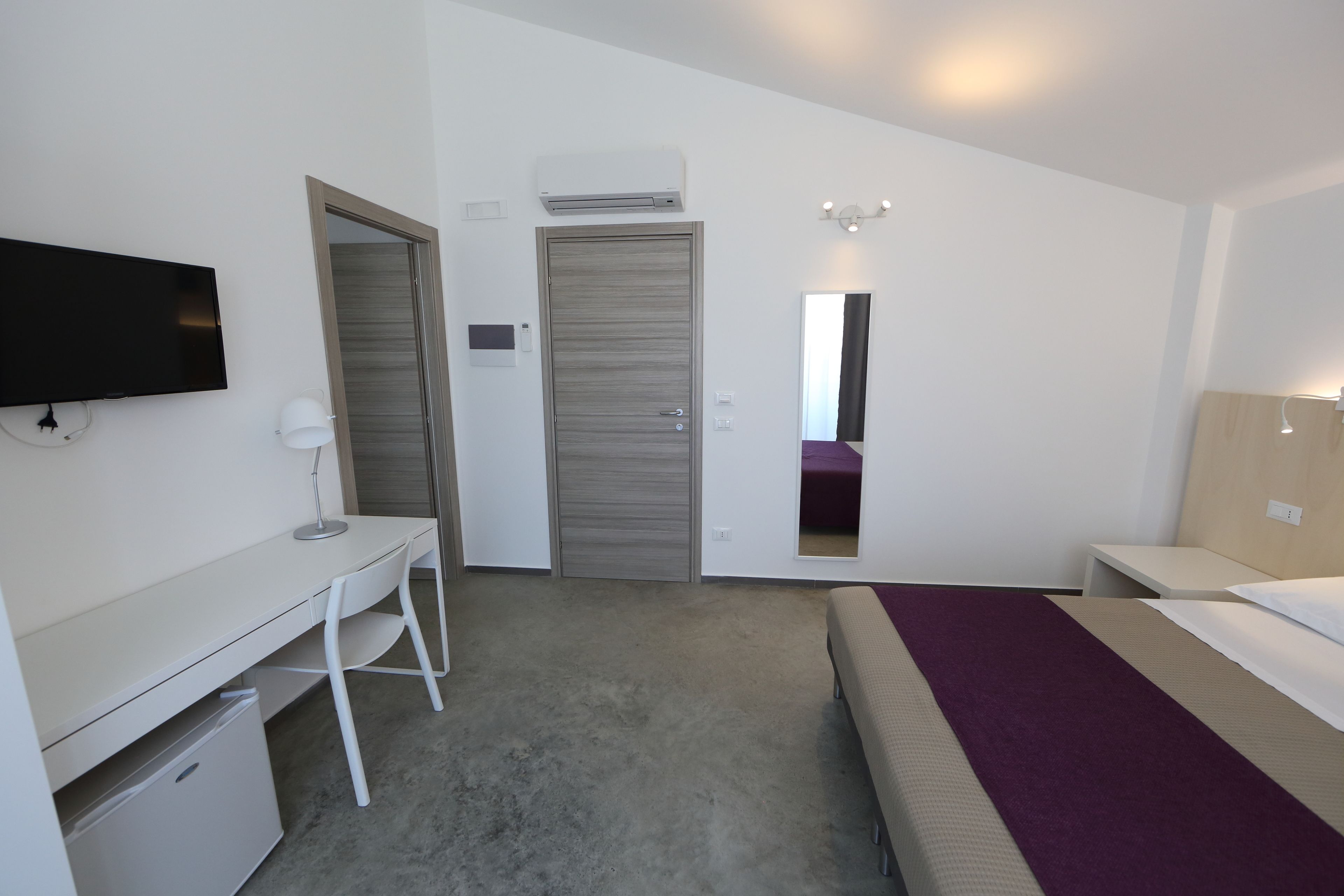 Una habitación de hotel con cama, escritorio, silla, televisor, armario y espejo.