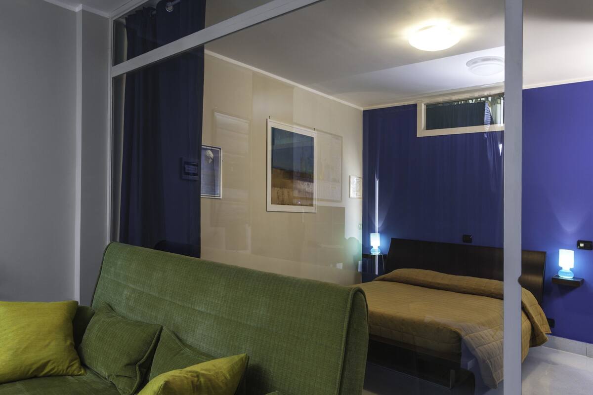 A hotel room with a green sofa, a bed with a headboard, a nightstand with a lamp, and a window with curtains.