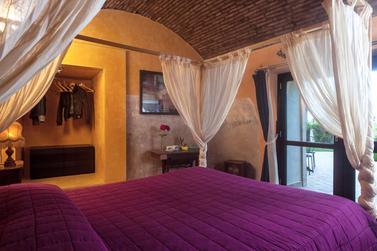 A bedroom with a large bed, a bedside table, a lamp, a wardrobe, and a window with curtains.
