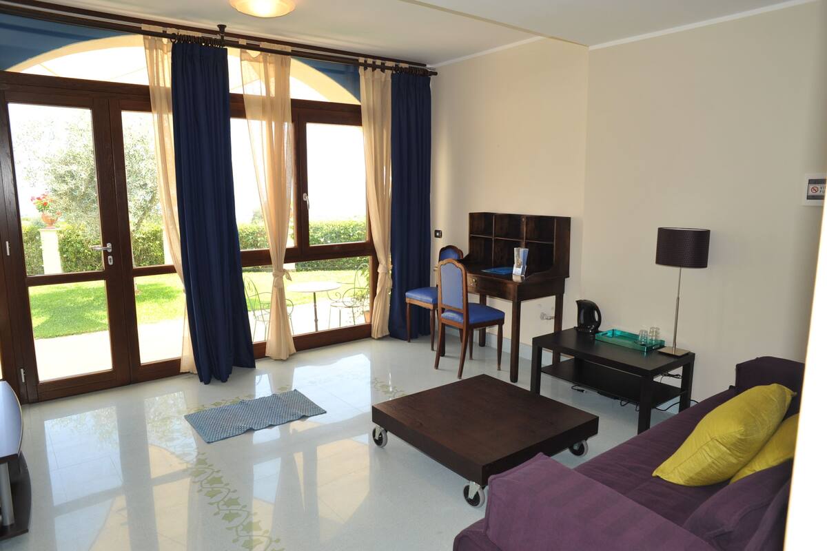 A room with a sofa, a desk with a chair, a coffee machine, and a TV. There is a sliding glass door leading to a balcony with a view of greenery.