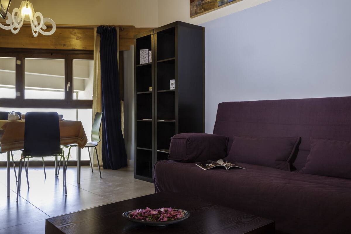A living room with a dark-colored sofa, a coffee table with a flower arrangement, a bookshelf, and a dining area with chairs and a table.