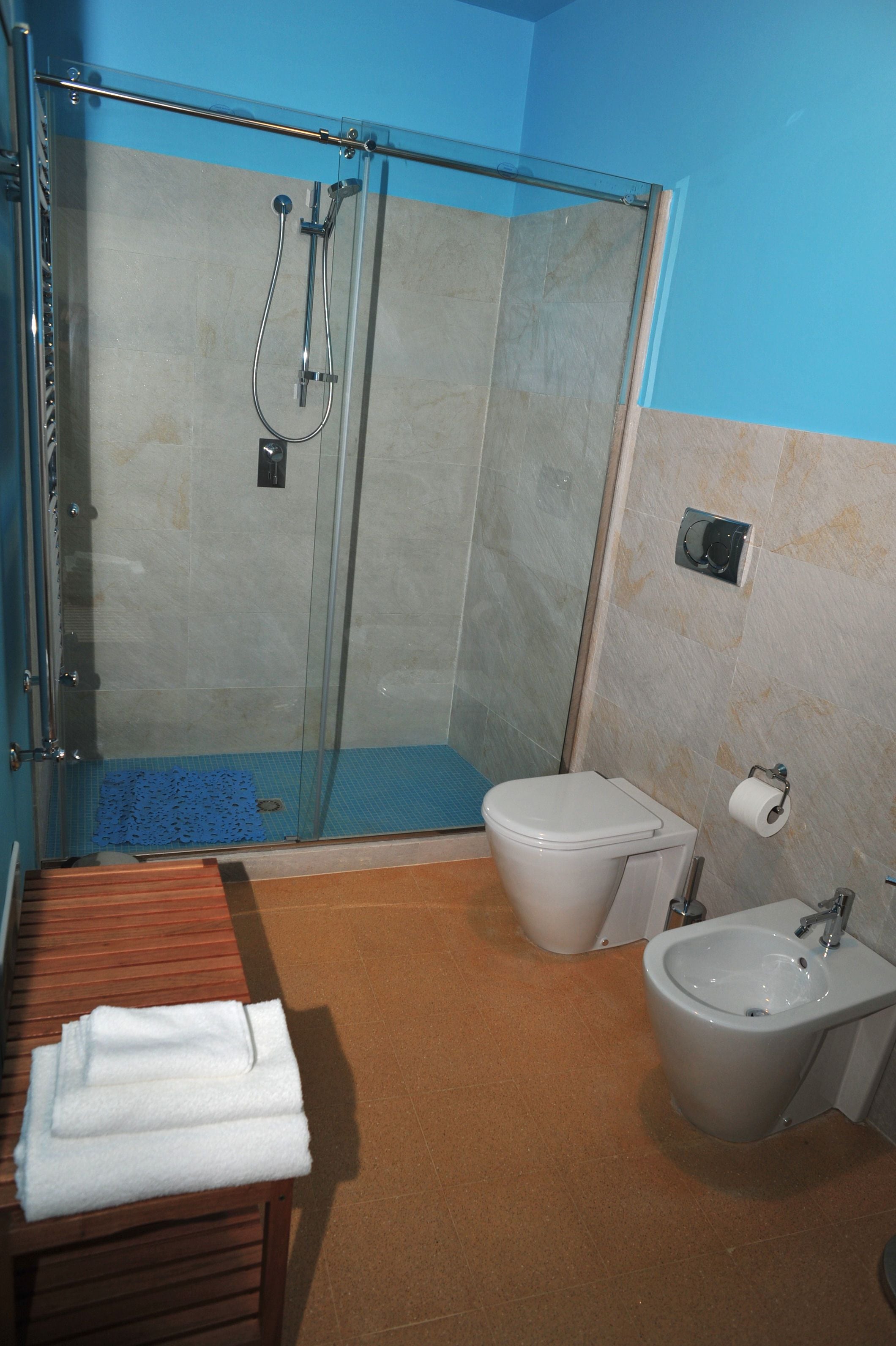 Baño con ducha de vidrio, inodoro, bidé y banco de madera con toallas.