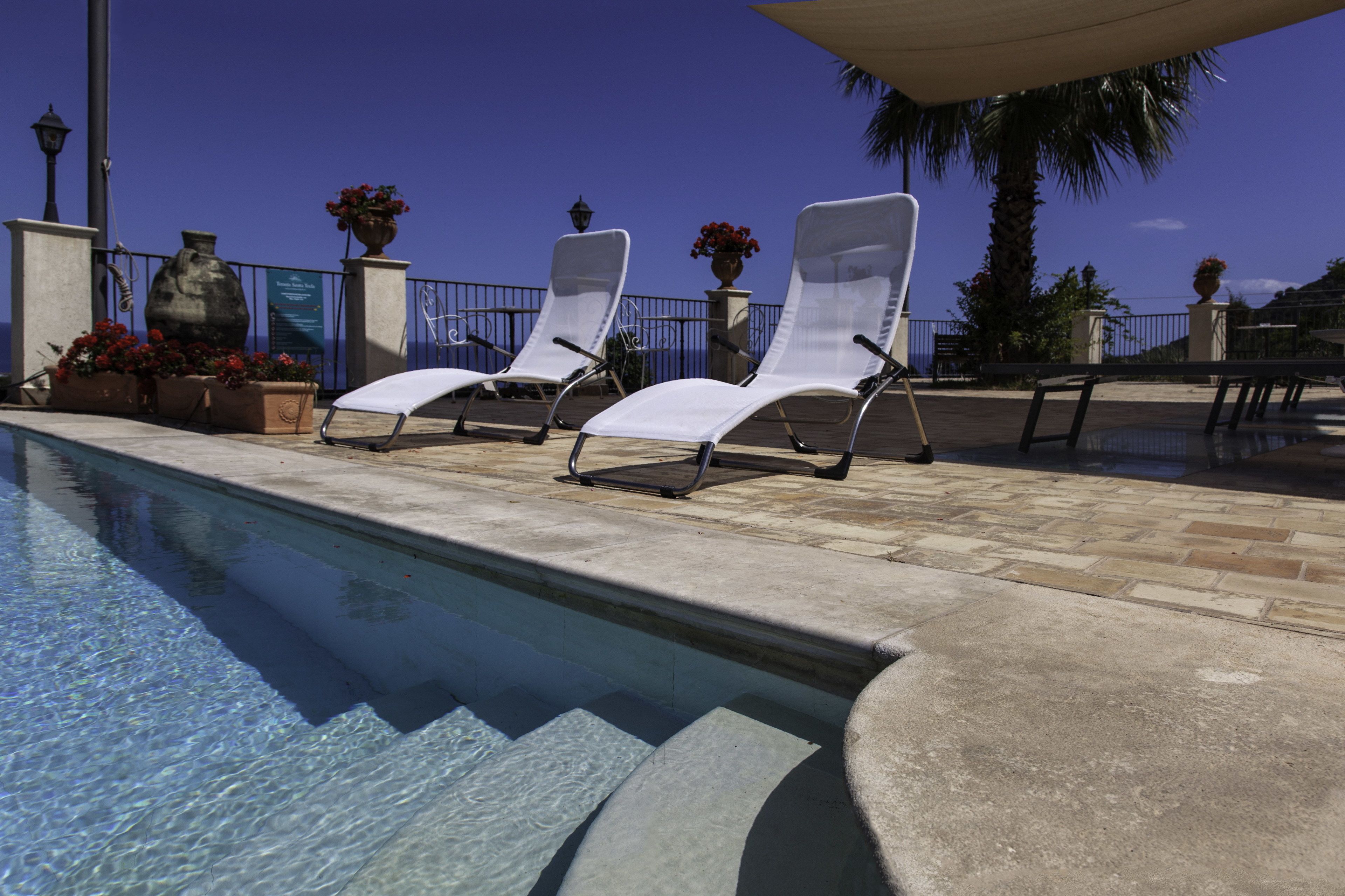 Uma área à beira da piscina com espreguiçadeiras, piscina de concreto e vasos decorativos com flores.