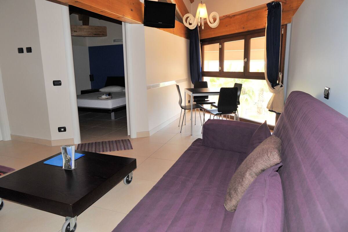 A room with a purple sofa, a small table with a book, a bed, a desk with a chair, and a television mounted on the wall.