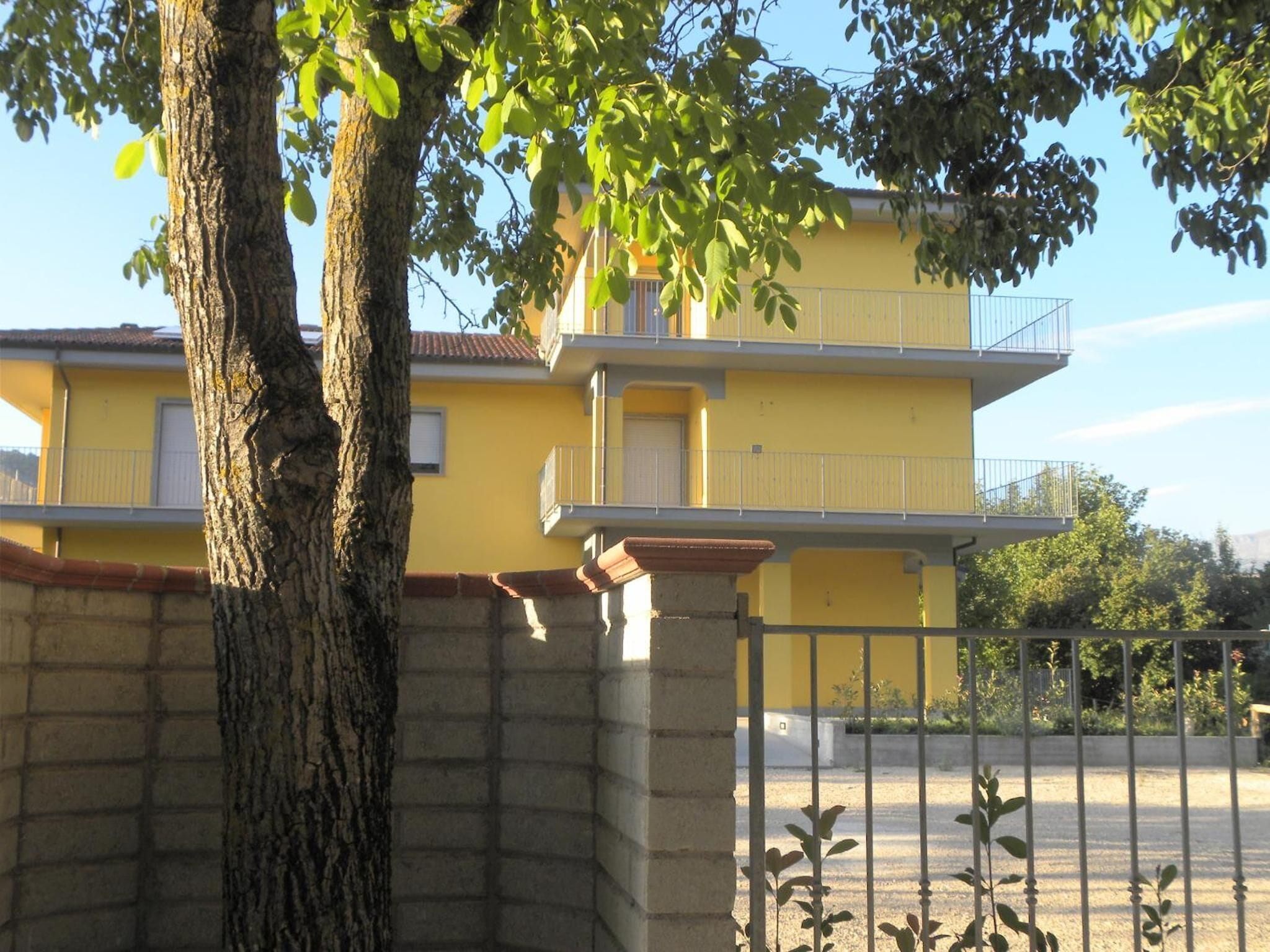 A yellow building with a balcony, a tree in the foreground, and a metal fence.