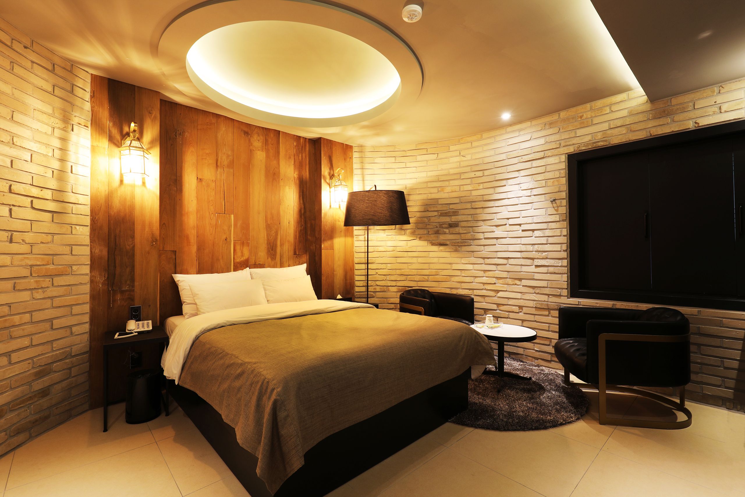 A modern hotel room with a brick wall, a large bed, a round ceiling light, a small table with a chair, and a TV mounted on the wall.