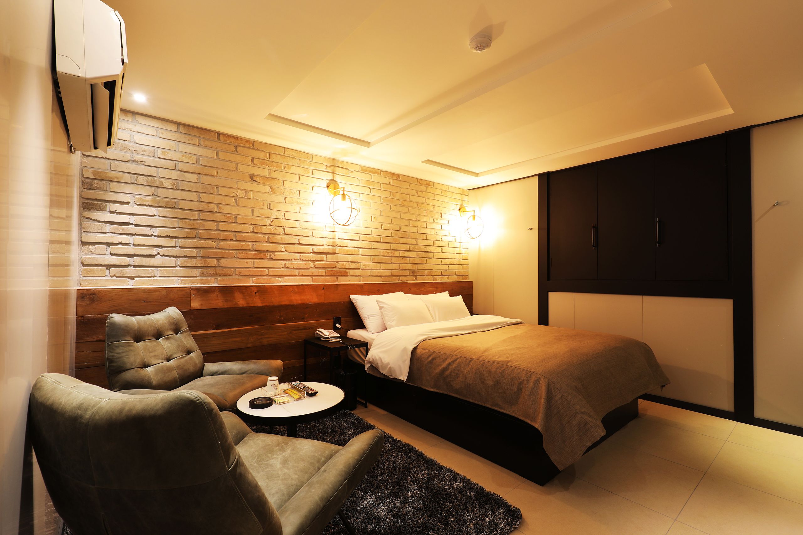 A modern hotel room with a brick accent wall, a bed with a headboard, a seating area with a round table, and a wardrobe.