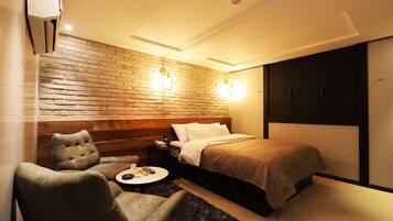 A modern hotel room with a brick accent wall, a bed with a headboard, a seating area with a round table, and a wardrobe.