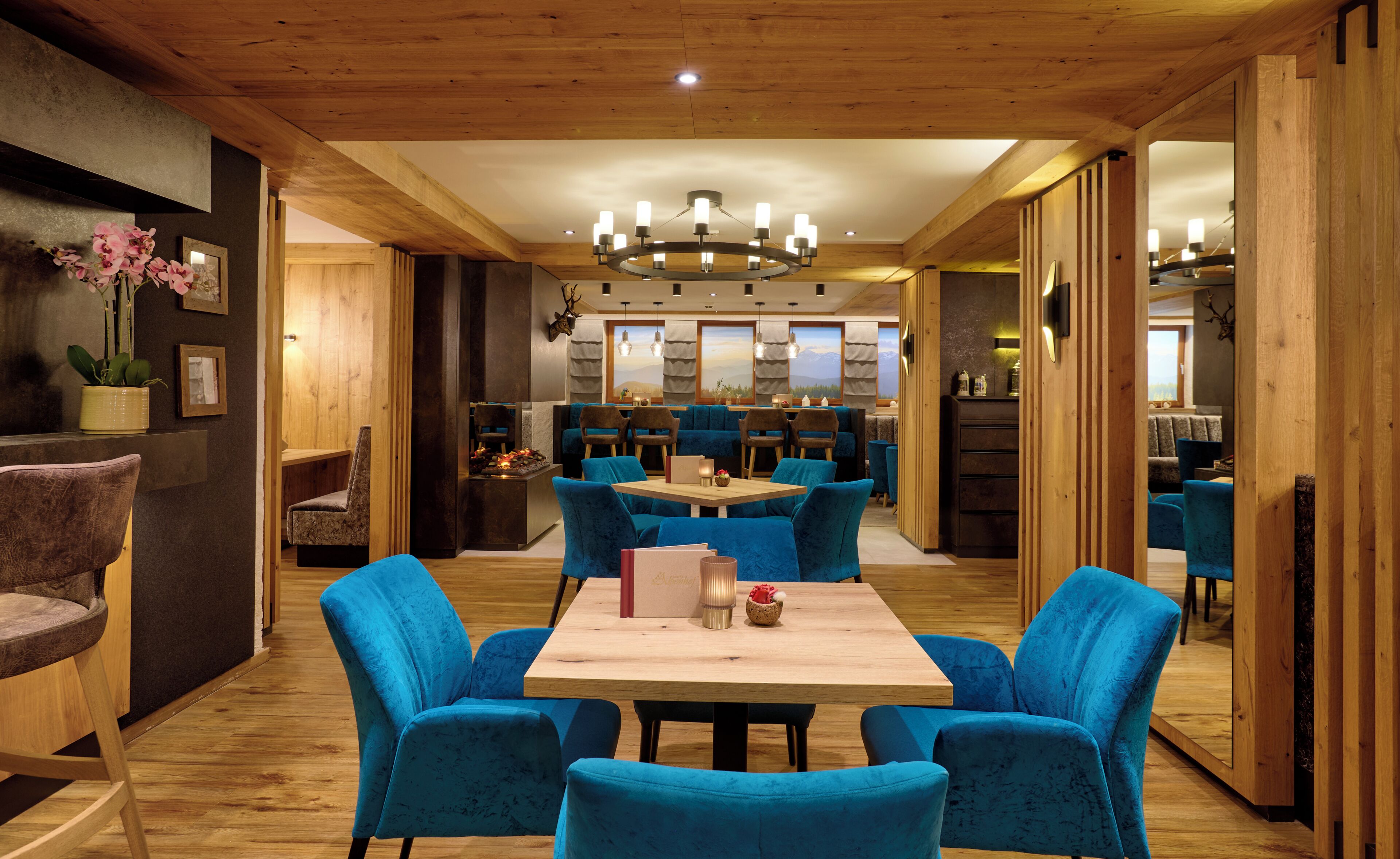 A modern interior with a wooden ceiling, a central dining table surrounded by blue chairs, and a kitchen area in the background.
