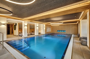 An indoor swimming pool with a wooden ceiling, steps leading into the pool, and a seating area with cushions. - Hotel Alpenhof (Bad Wiessee)