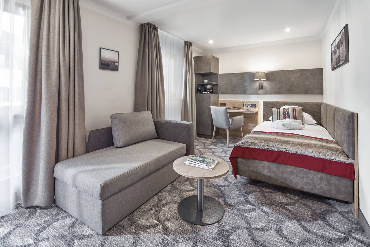 Ein Hotelzimmer mit einer Couch, einem kleinen runden Tisch, einem Bett mit roter und weißer Decke, einem Schreibtisch mit Stuhl und einer Kaffeemaschine.