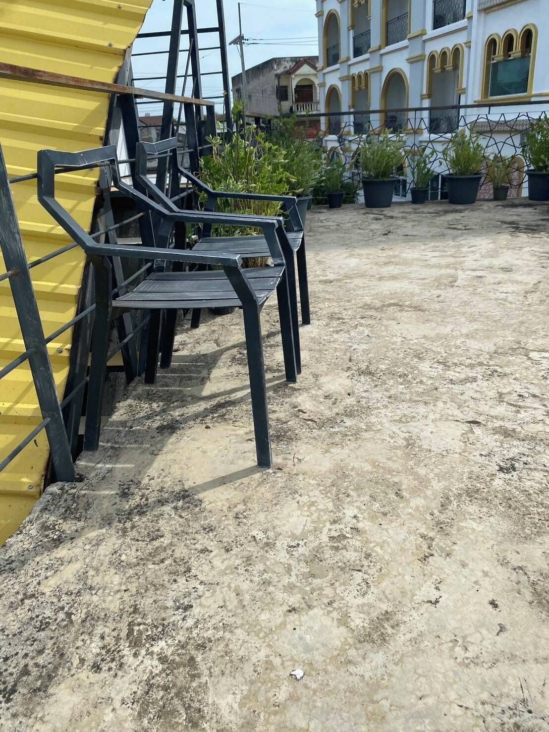 Bancos de metal al aire libre, una estructura amarilla y plantas en macetas en un patio con un edificio al fondo.