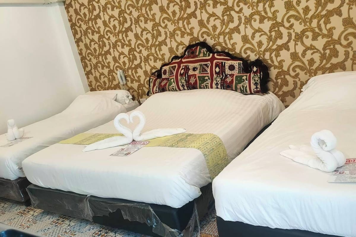 Three single beds with white linens, each with a towel and a small card. The room has patterned wallpaper and a wooden headboard.