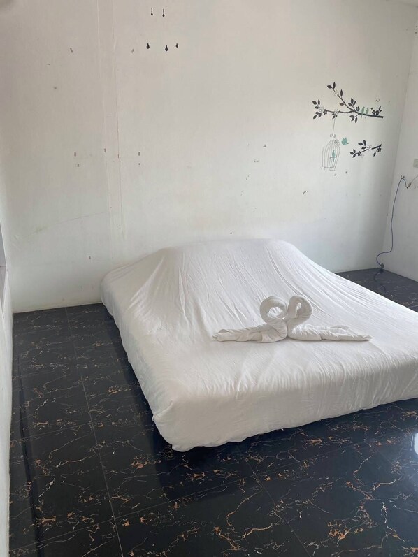 Una cama individual con un colchón blanco y una toalla con forma de cisne, en una habitación con suelo de mármol oscuro y una pared blanca decorada con una rama y un pájaro.