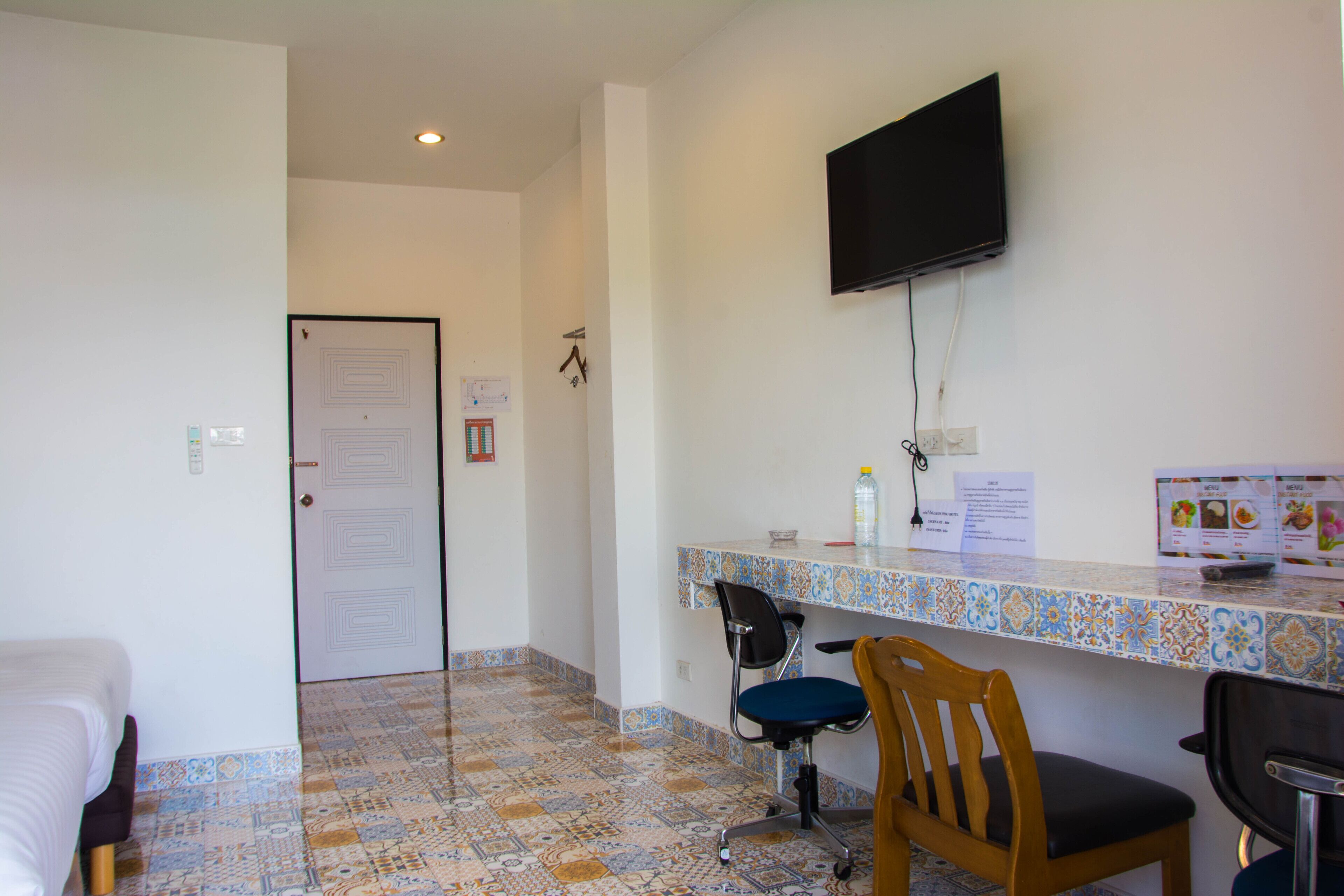 A hotel room with a tiled floor, a bed, a desk with a chair, a television mounted on the wall, and a door with a sign.