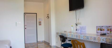 A hotel room with a tiled floor, a bed, a desk with a chair, a television mounted on the wall, and a door with a sign.
