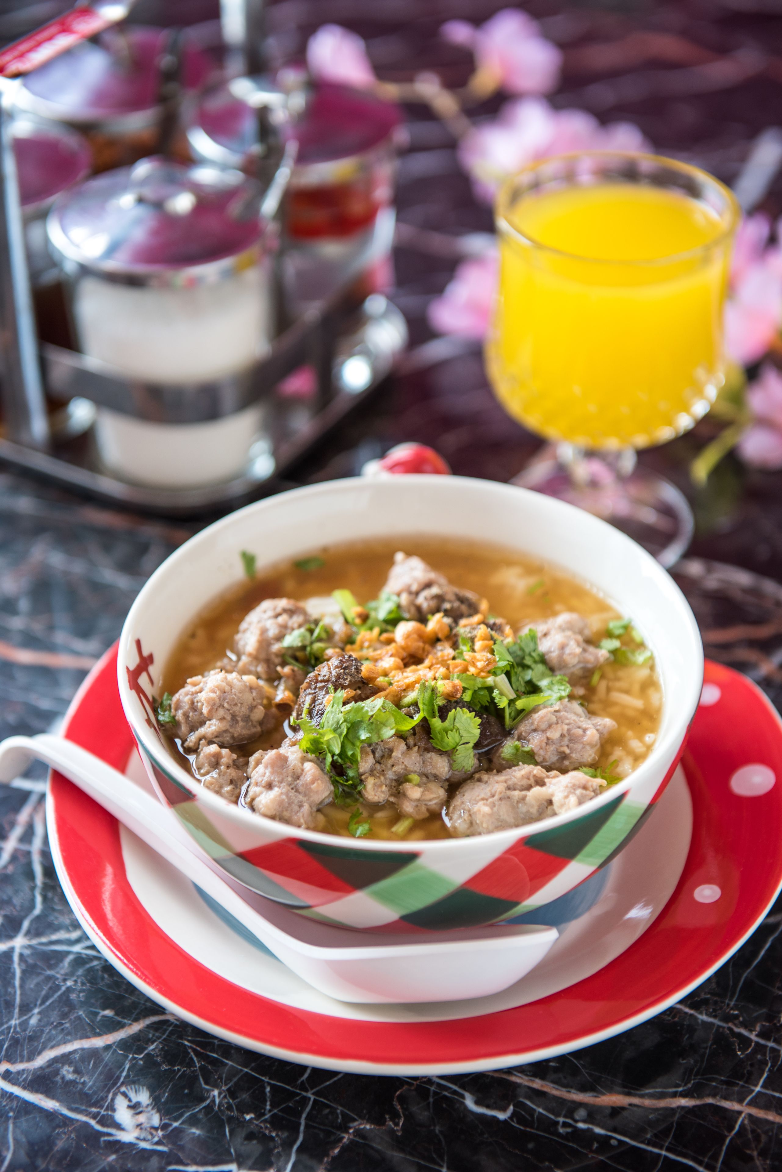 Un tazón de sopa con albóndigas, adornado con hierbas, sobre un plato con diseño rojo y blanco.
