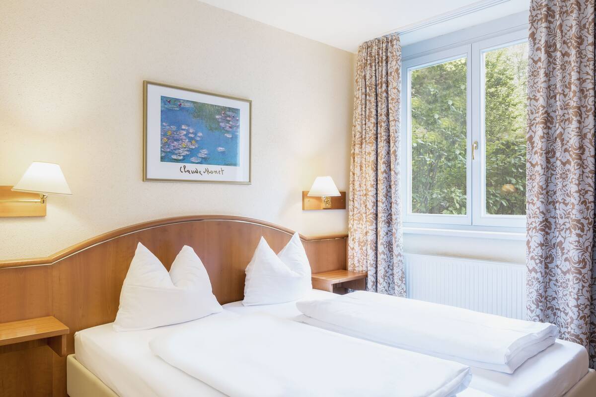 A hotel room with a double bed, two bedside lamps, a framed painting, and a window with curtains.