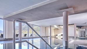 Un área de piscina interior moderna con paredes de vidrio transparente, barandales metálicos y vistas a las montañas.