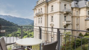 Un balcón con sillas de mimbre y una mesa con vistas a un paisaje montañoso y un edificio grande de arquitectura clásica.